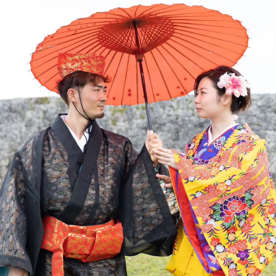 赤い和傘をさし沖縄の伝統衣装を着て見つめ合う若い夫婦